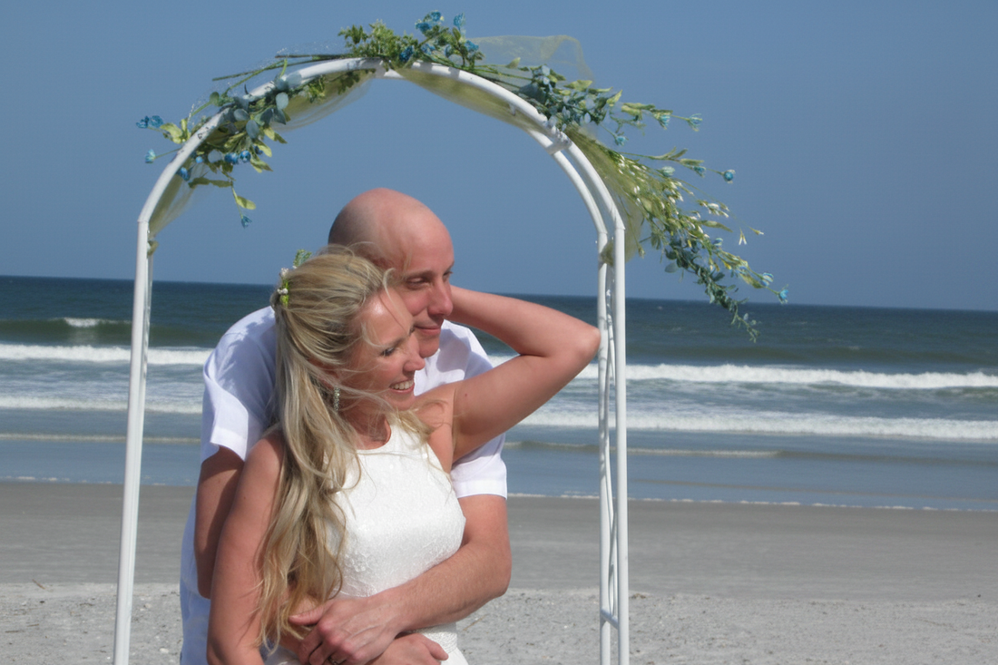 Jana and Geoff Yockey, founders of Garlic Gods, embracing under a white floral arch on Jacksonville Beach Florida with Atlantic Ocean waves and blue sky behind them
